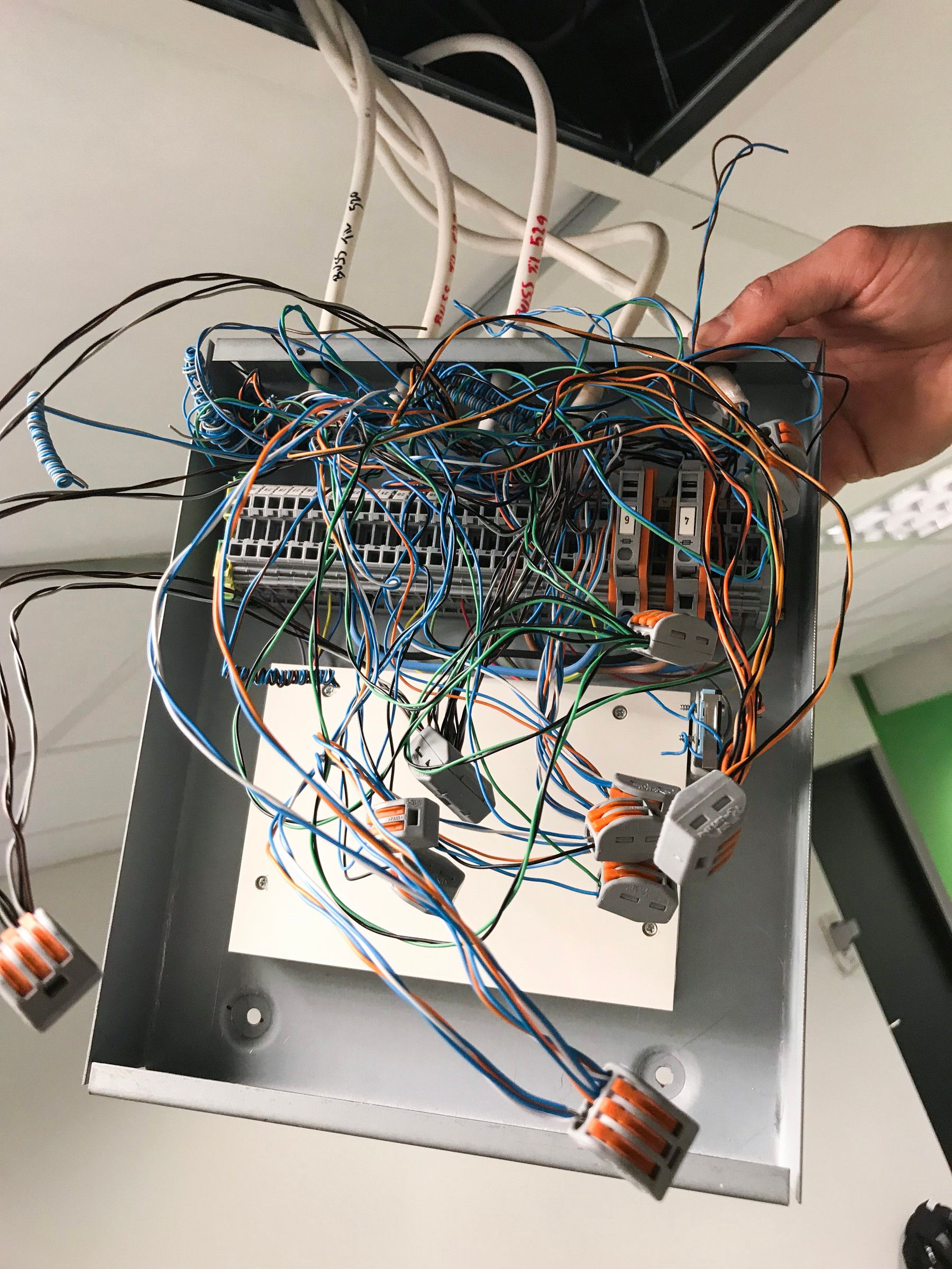 Open wiring box under the ceiling with a tangle of loose, coloured wires and terminals, held up by a hand
